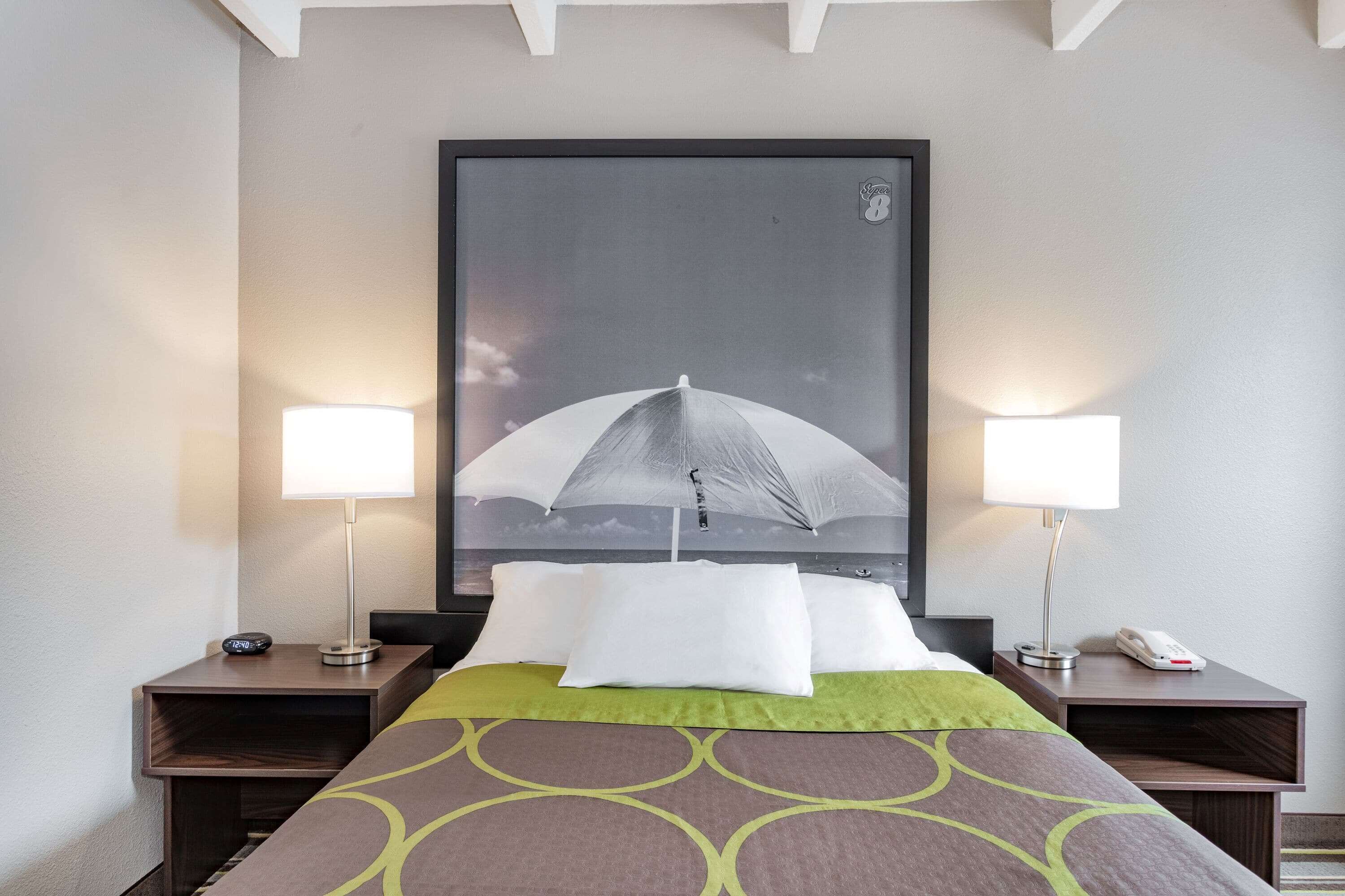 A hotel room with a bed, two bedside tables, two lamps, a large framed picture on the wall, and a green throw blanket.