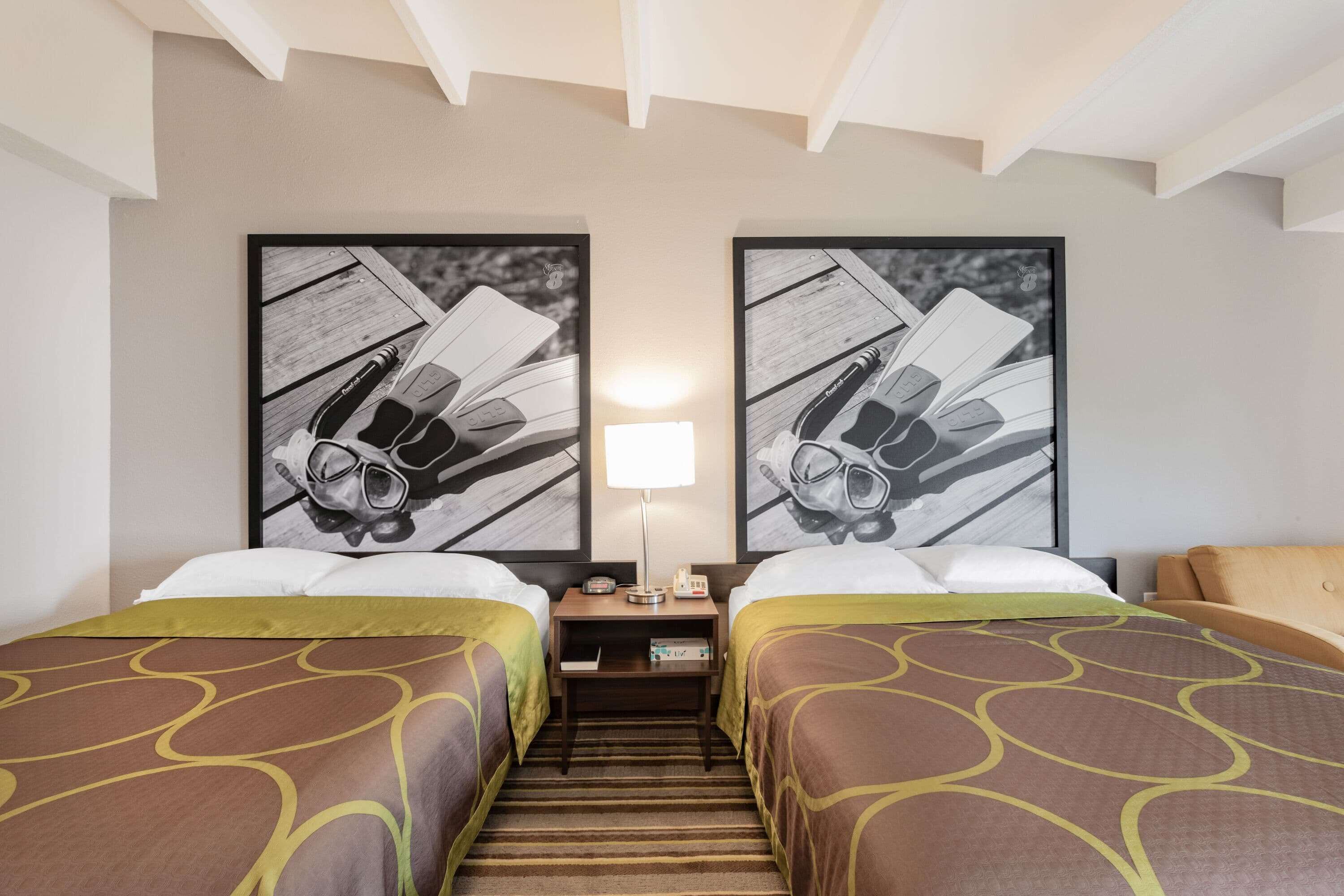 A hotel room with two beds, a nightstand, a lamp, and two framed pictures on the wall.