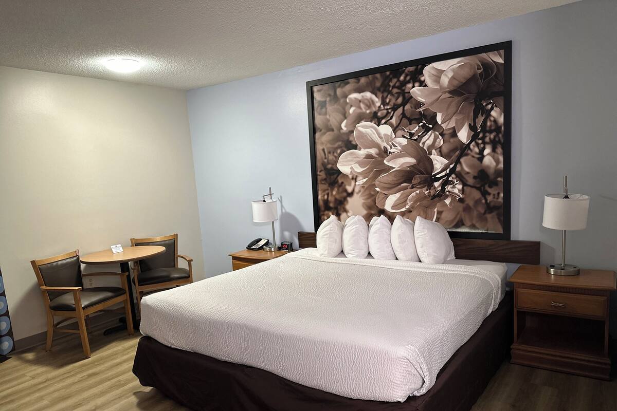 A hotel room with a large bed, a desk with a chair, a nightstand with a lamp, and a wall-mounted artwork.