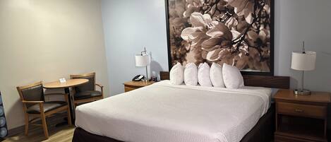 A hotel room with a large bed, a desk with a chair, a nightstand with a lamp, and a wall-mounted artwork.