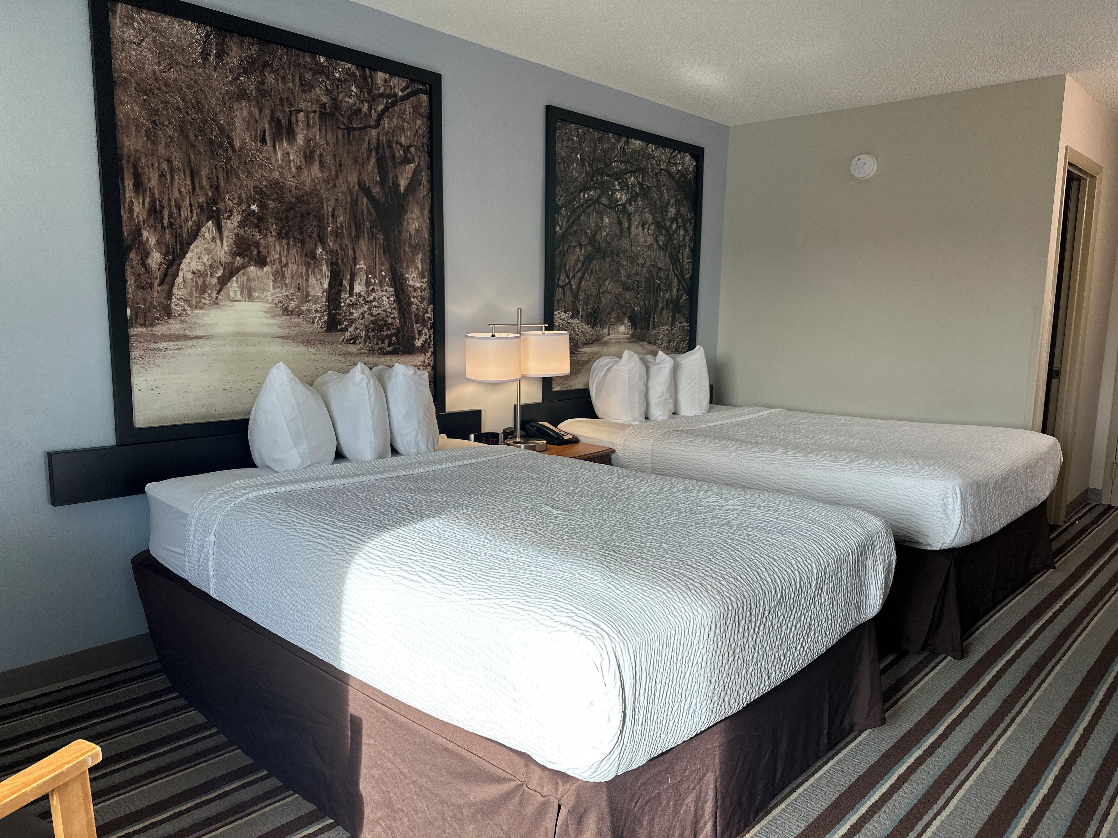 A hotel room with two beds, a nightstand with a lamp, and large framed pictures on the wall.