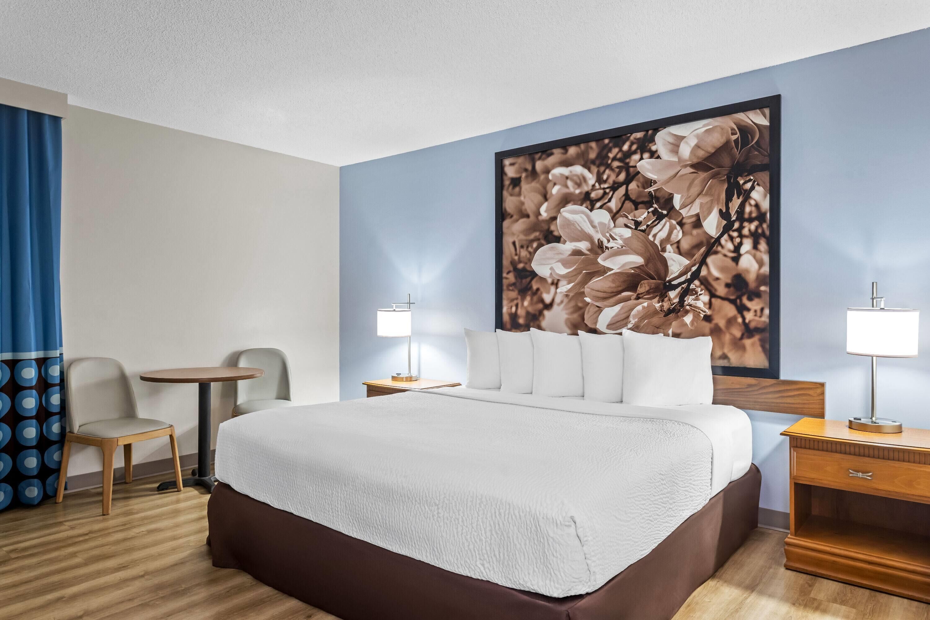 A hotel room with a large bed, two bedside tables, a small round table, a chair, and a large floral artwork on the wall.