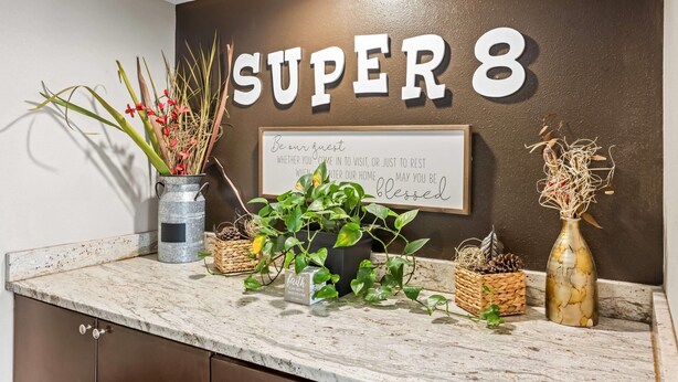 Una zona de recepción con un cartel que dice "SUPER 8" y un mensaje de bienvenida en la pared. Hay plantas en macetas y objetos decorativos sobre el mostrador.