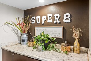 A reception area with a sign reading "SUPER 8" and a welcome message on the wall. There are potted plants and decorative items on the counter. - Super 8 by Wyndham Valdosta GA I-75 (Valdosta)