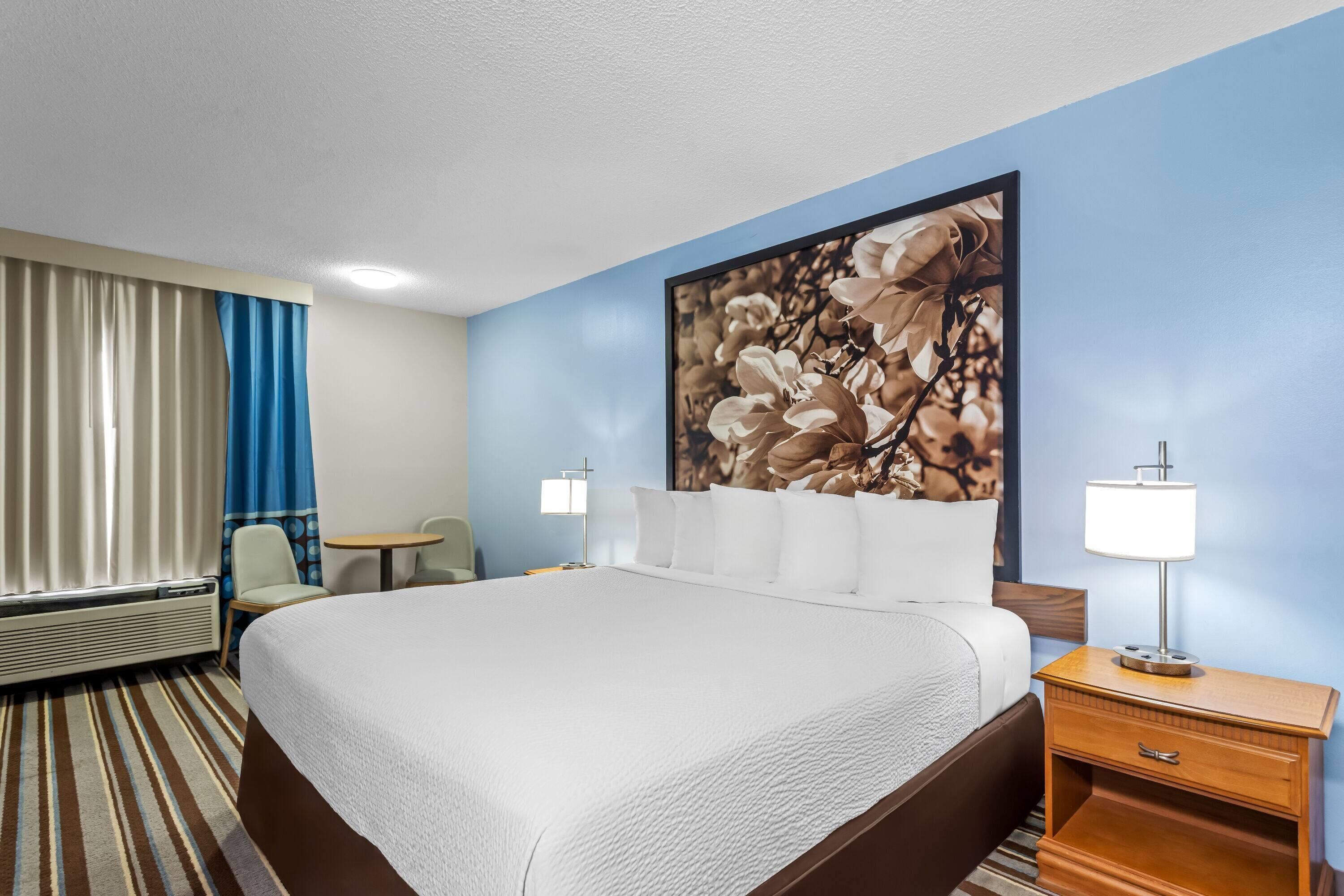 Una habitación de hotel con una cama grande, una mesita de noche con lámpara, una silla, una pequeña mesa y un aire acondicionado instalado en la pared.