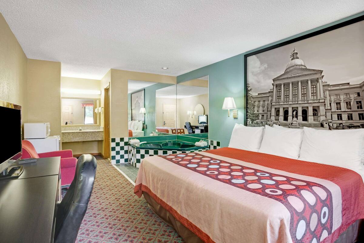 Camera d'albergo con un letto, una scrivania, una sedia, una televisione, un bagno con lavandino e specchio, e un'ampia finestra con vista su un edificio.