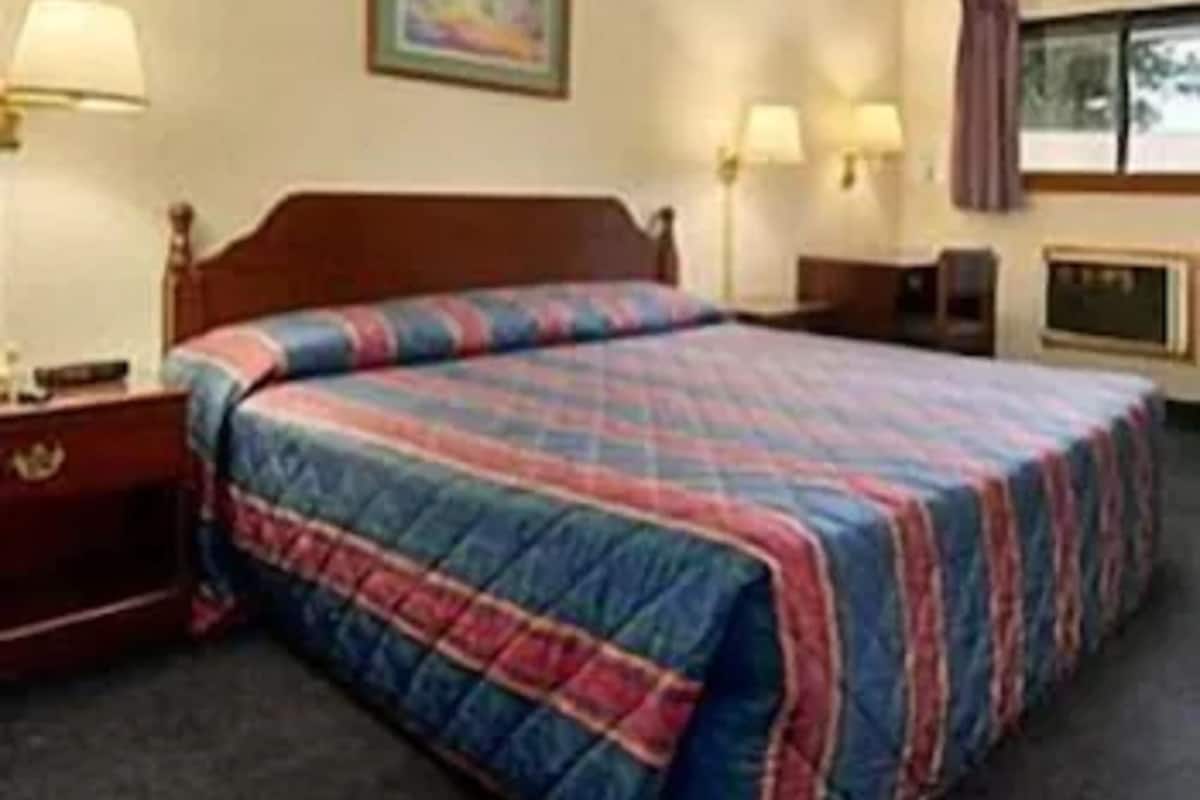 A hotel room with a bed, a nightstand, a lamp, a painting on the wall, and a window with curtains.