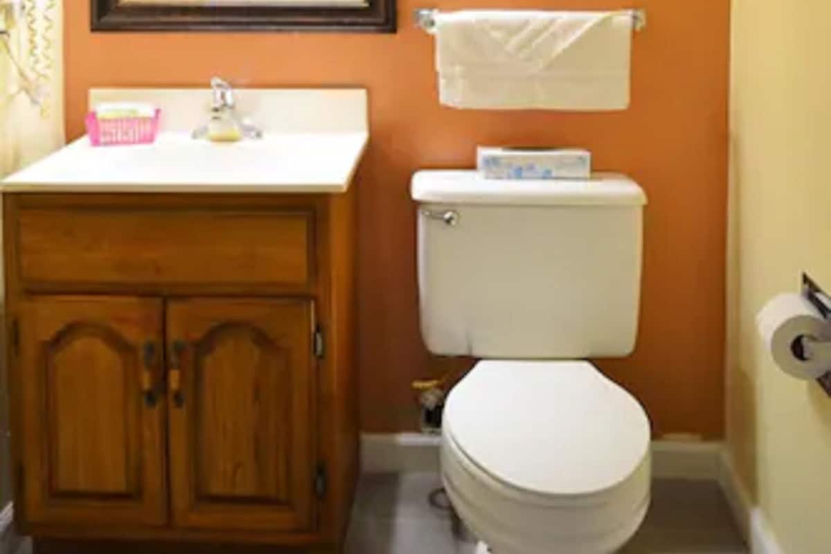 A bathroom with an orange wall, a wooden vanity with a mirror above it, a white toilet, and a towel hanging on a rack.
