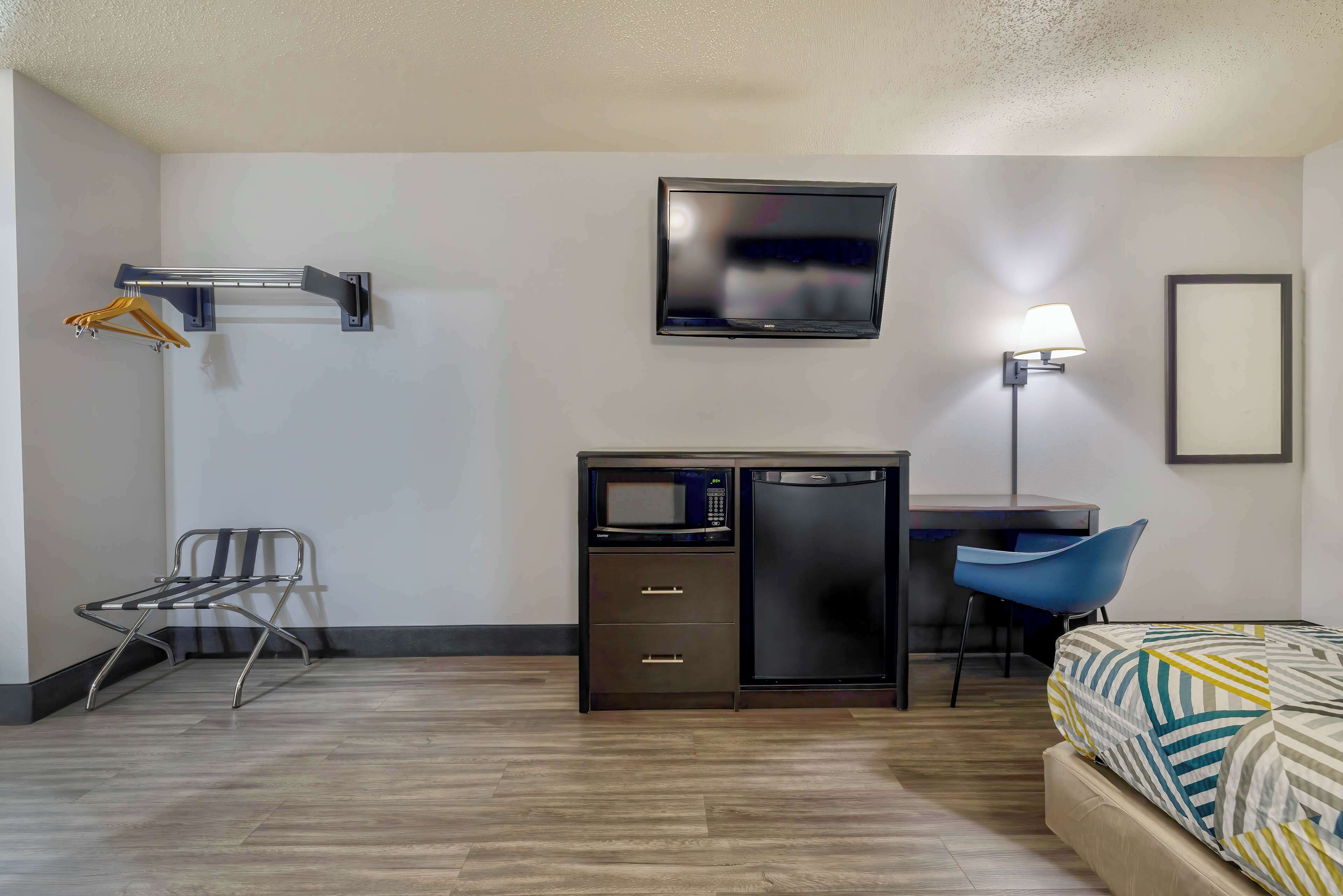 A hotel room with a bed, a desk with a microwave, a TV, a chair, and a coat rack.