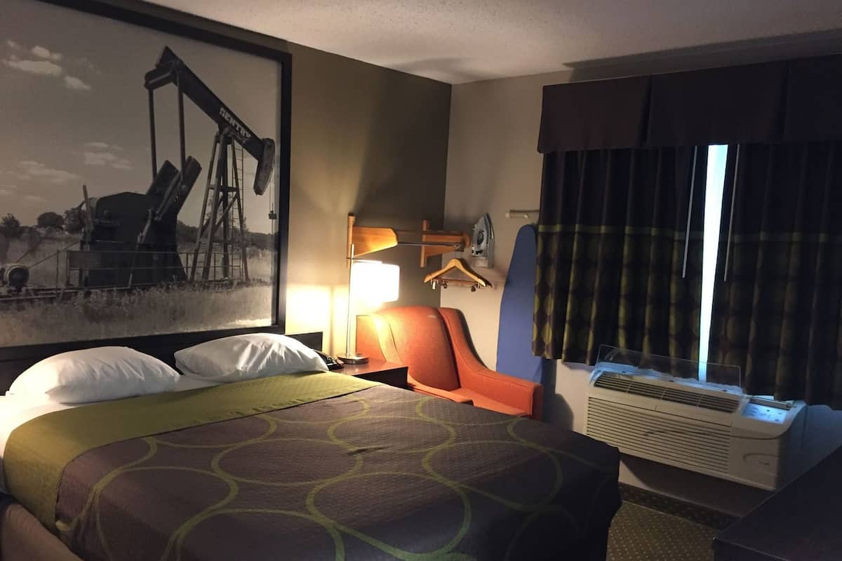 Ein Hotelzimmer mit einem Bett, einem Schreibtisch, einem Stuhl, einem Fenster mit Vorhängen, einer Wand-Klimaanlage und einem Gemälde einer Ölplattform.