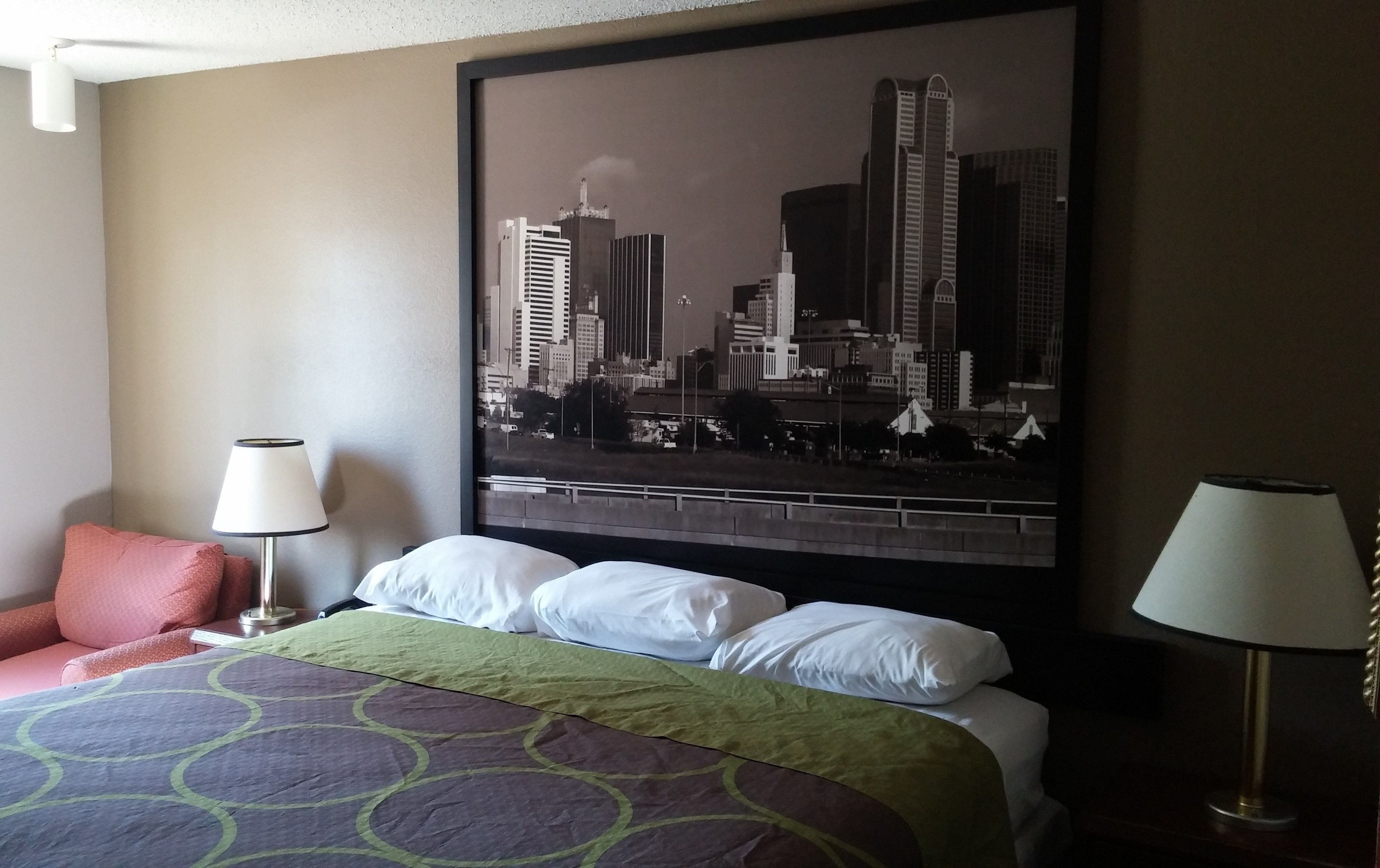 Habitación de hotel con cama, un cuadro de paisaje urbano en la pared, una silla rosa y dos lámparas.