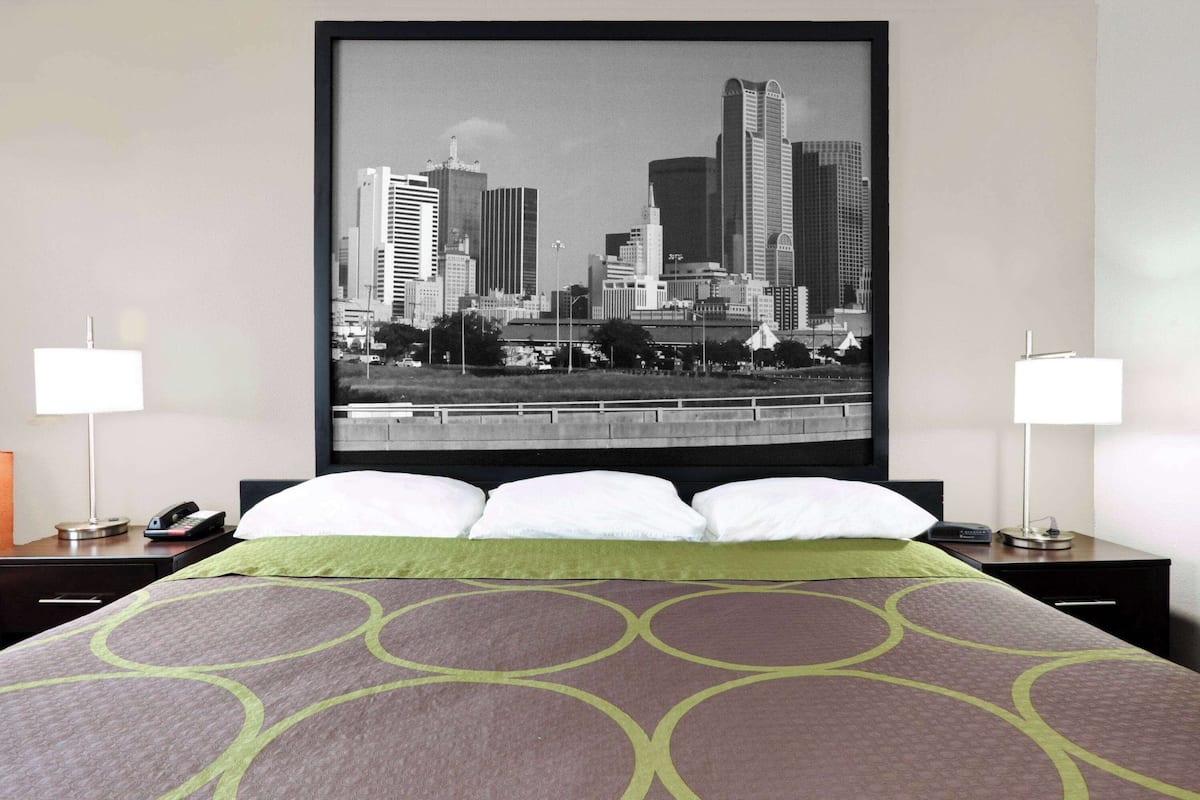 Quarto de hotel com uma cama grande, duas mesinhas de cabeceira, uma pintura urbana na parede e um edredom com padrão verde e marrom.