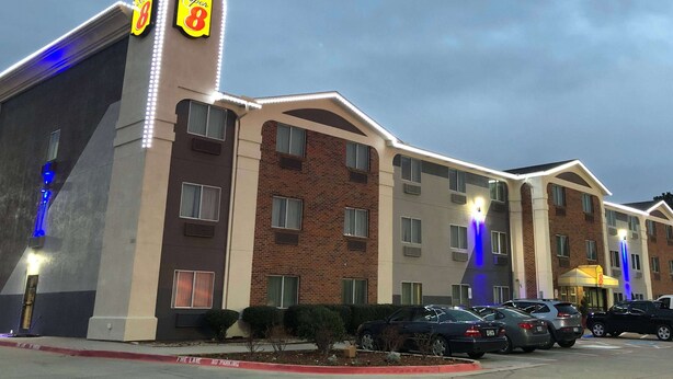 A hotel with a brick facade, multiple windows, and a parking lot with several cars. - Super 8 by Wyndham Bedford DFW Airport West (Bedford)