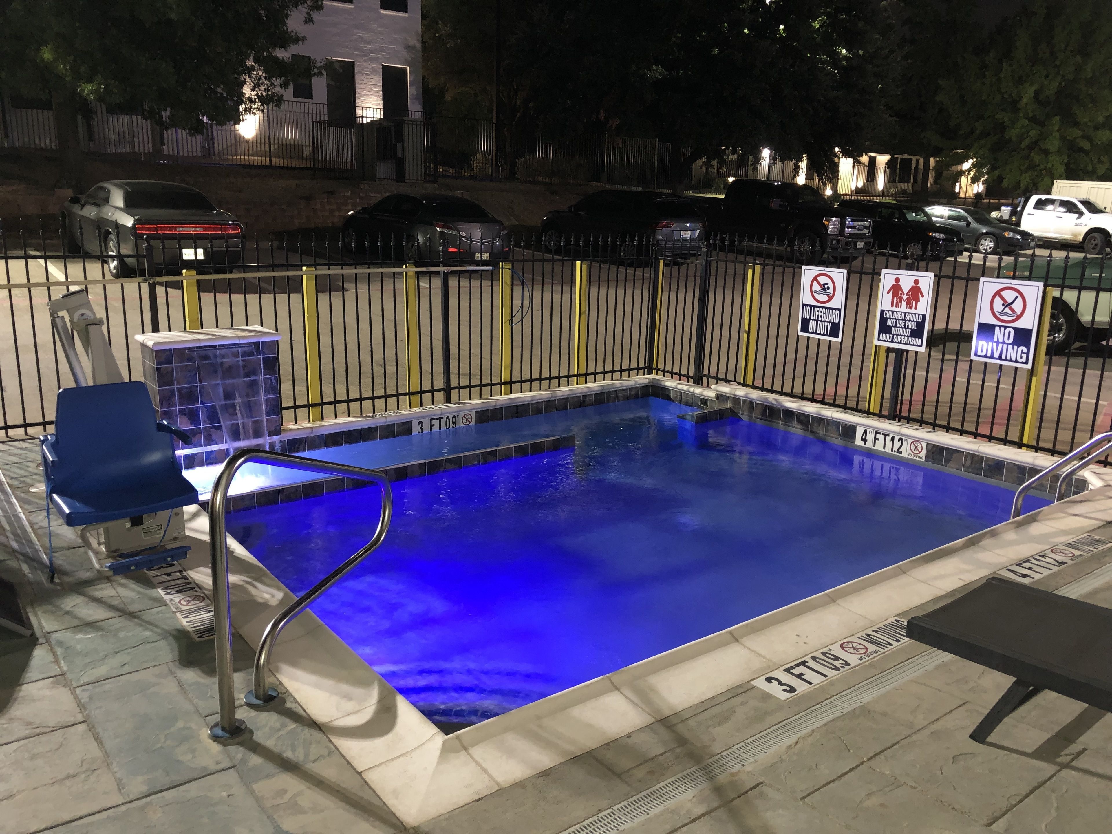An outdoor swimming pool with a blue tint, surrounded by a metal fence, and marked with safety signs.