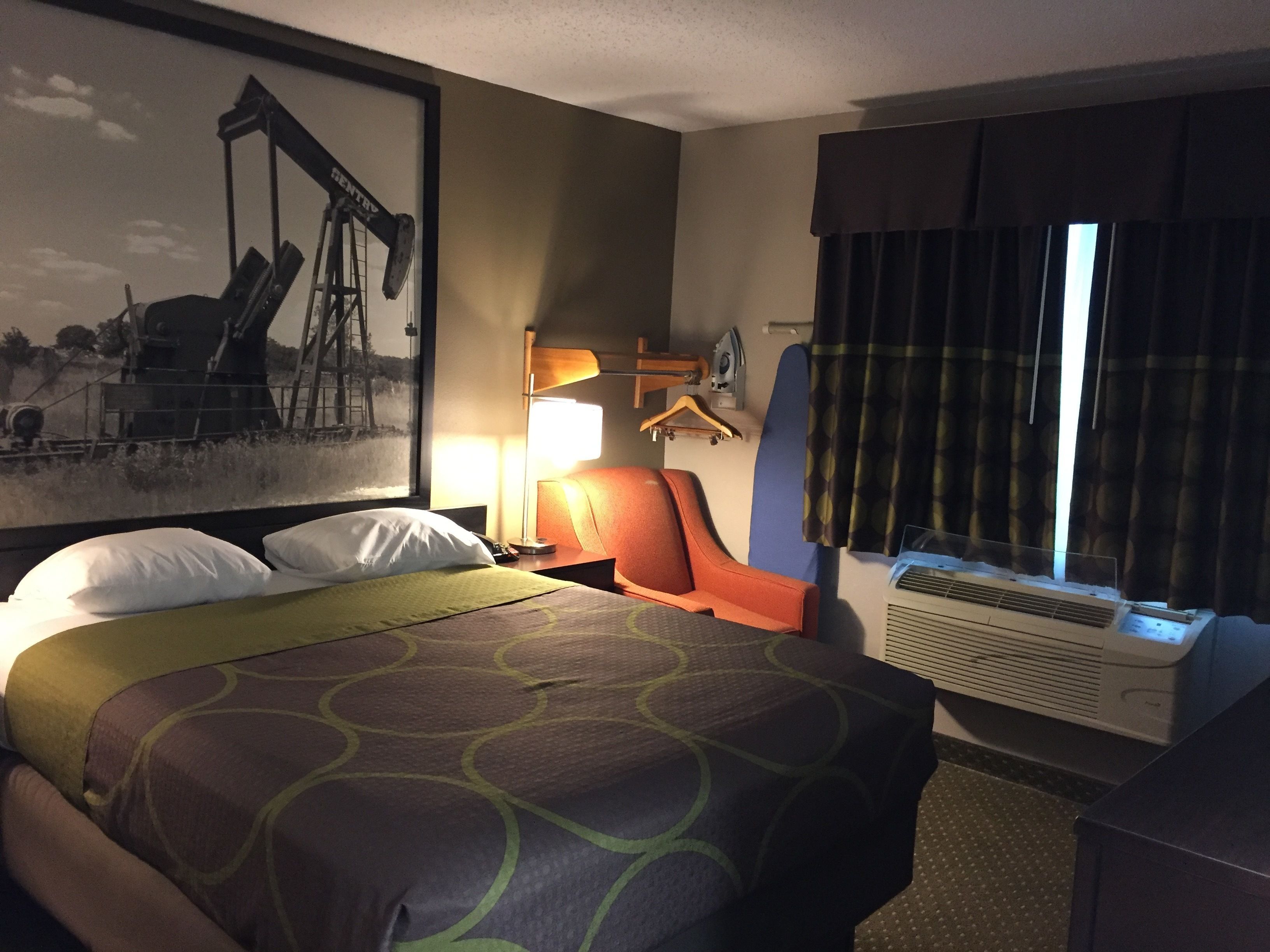 A hotel room with a bed, a desk, a chair, a window with curtains, an air conditioning unit, and a painting on the wall.