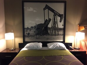 A hotel room with a bed, two bedside lamps, a framed picture of an oil pumpjack, and a telephone on the nightstand. - Super 8 by Wyndham Bedford DFW Airport West (Bedford)
