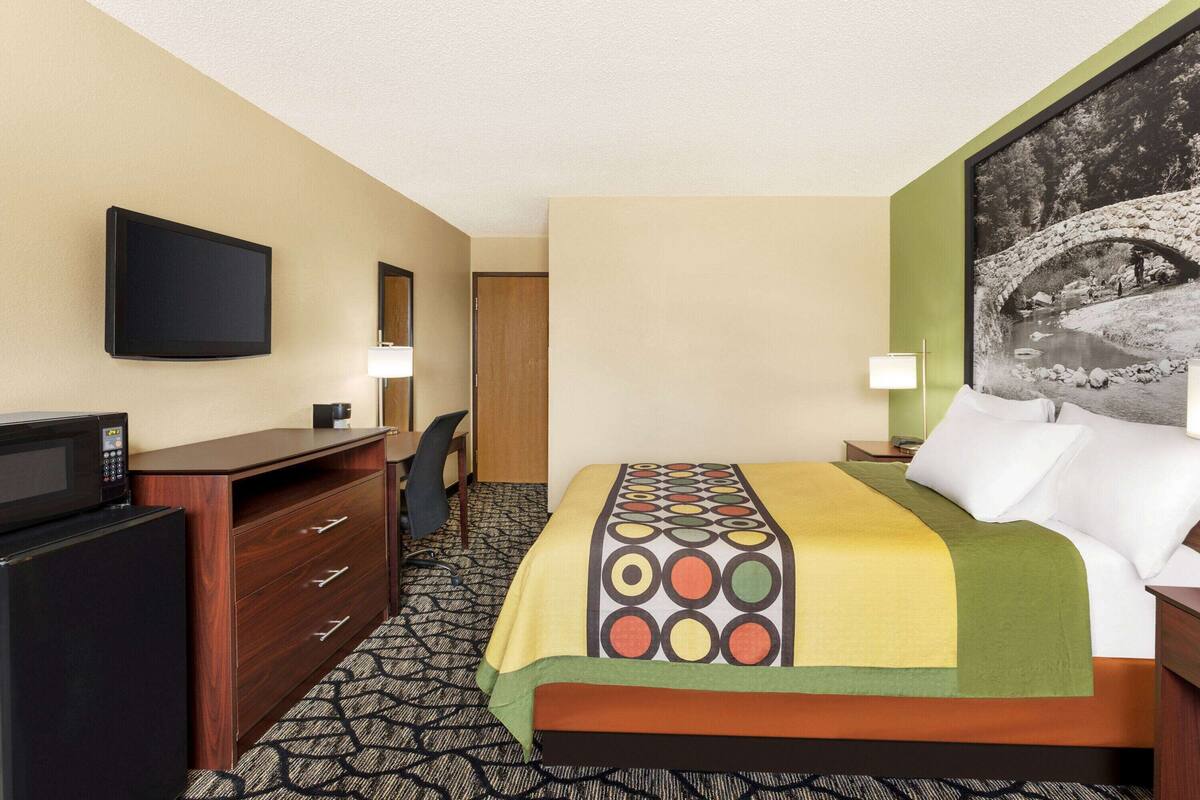 A hotel room with a bed, a desk, a chair, a TV, a microwave, and a painting on the wall.