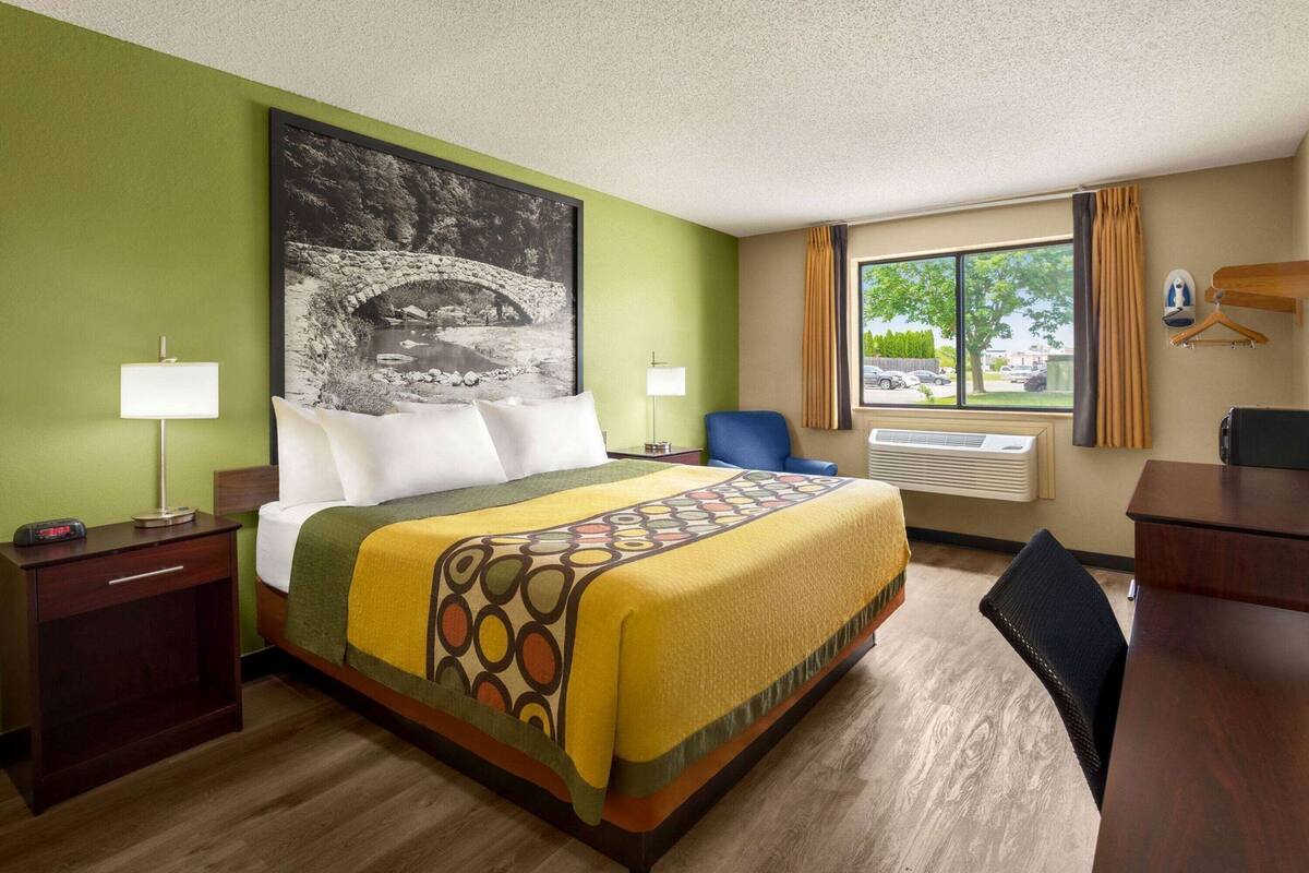 A hotel room with a bed, a nightstand, a desk, a chair, a window with curtains, and a wall-mounted heater.