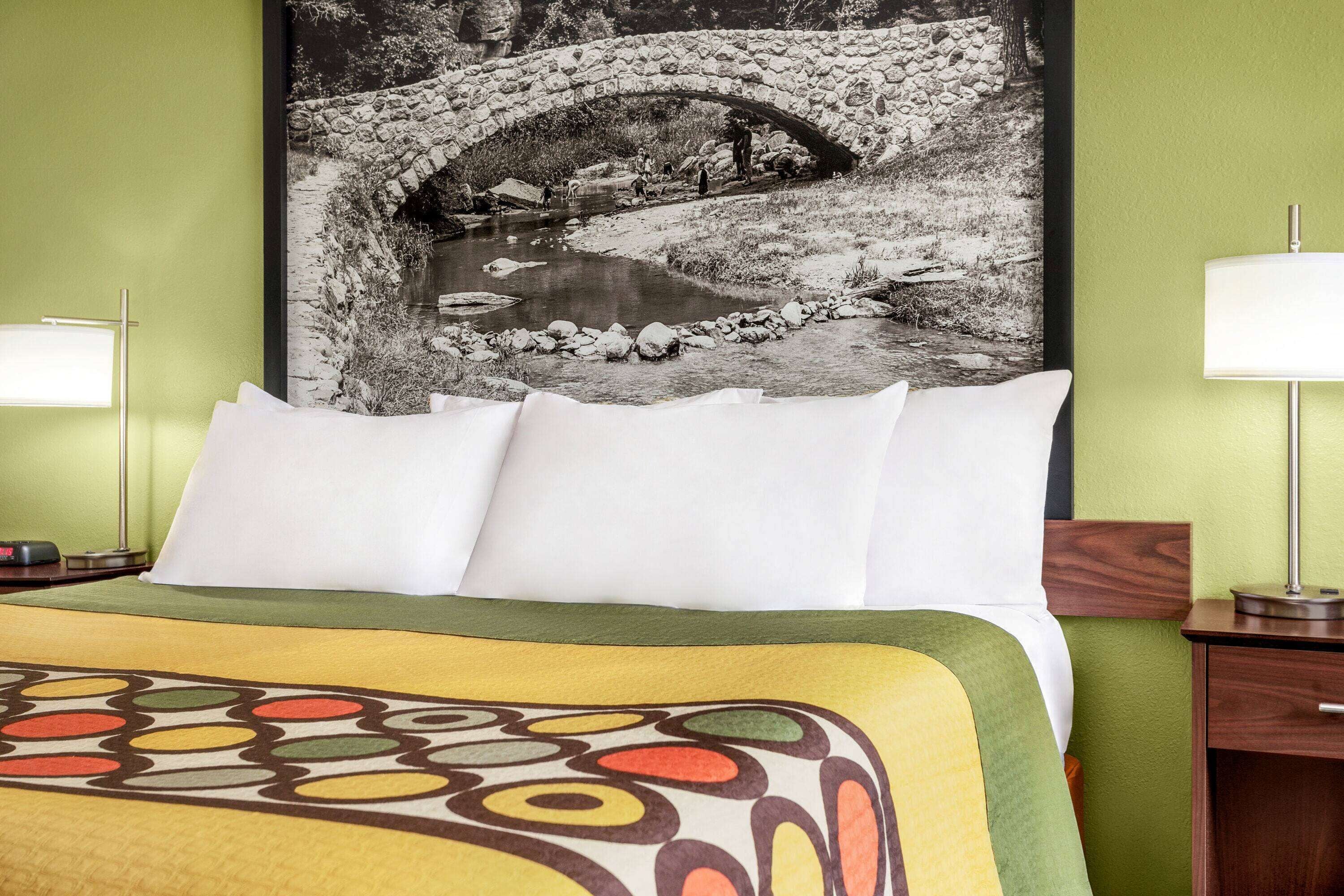 A neatly made bed with a colorful blanket, a black and white photo of a stone bridge over a river, and a lamp on a bedside table.