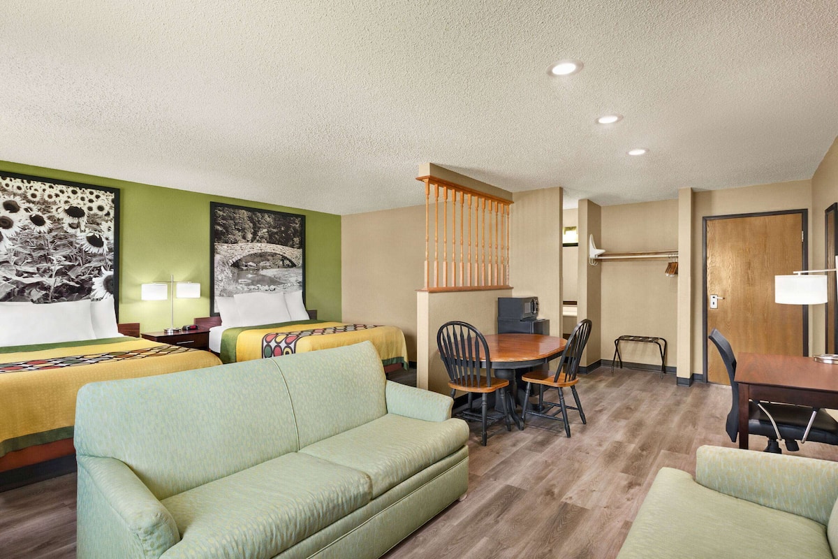 A hotel room with two beds, a green sofa, a dining table with chairs, and a small kitchen area.