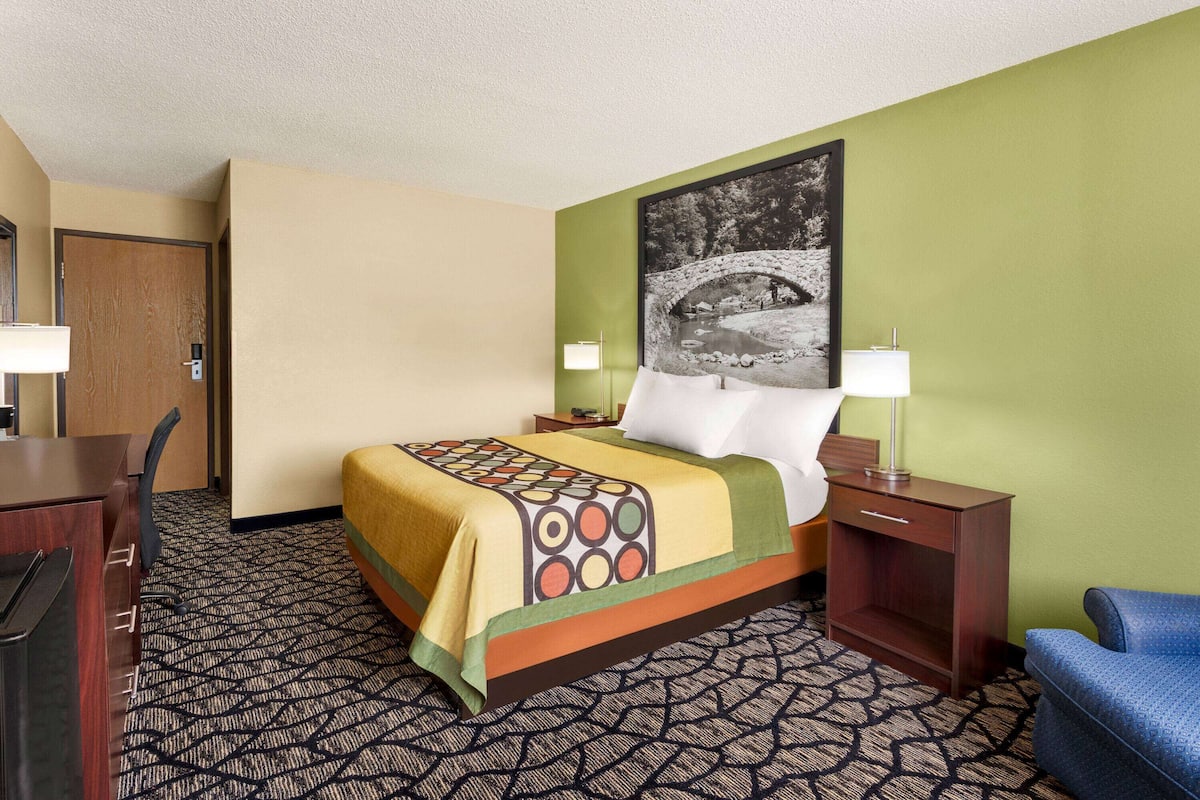 A hotel room with a bed, a desk, a chair, a nightstand, and a wall-mounted lamp.