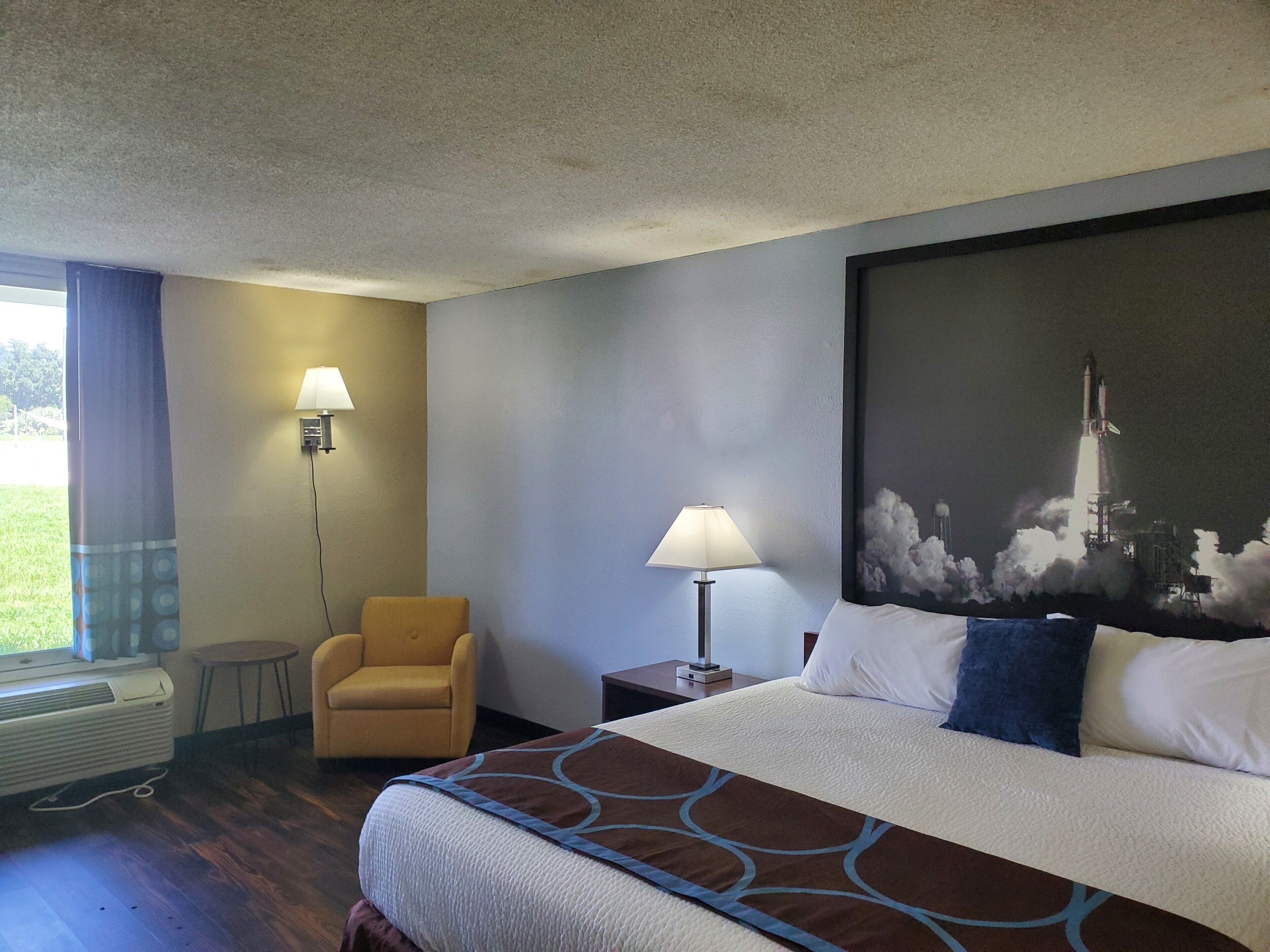 Une chambre d’hôtel avec un lit, deux lampes de chevet, une chaise, une petite table et un grand tableau représentant le lancement d’une fusée.