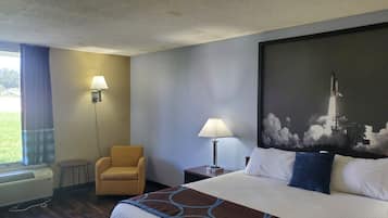Une chambre d’hôtel avec un lit, deux lampes de chevet, une chaise, une petite table et un grand tableau représentant le lancement d’une fusée.