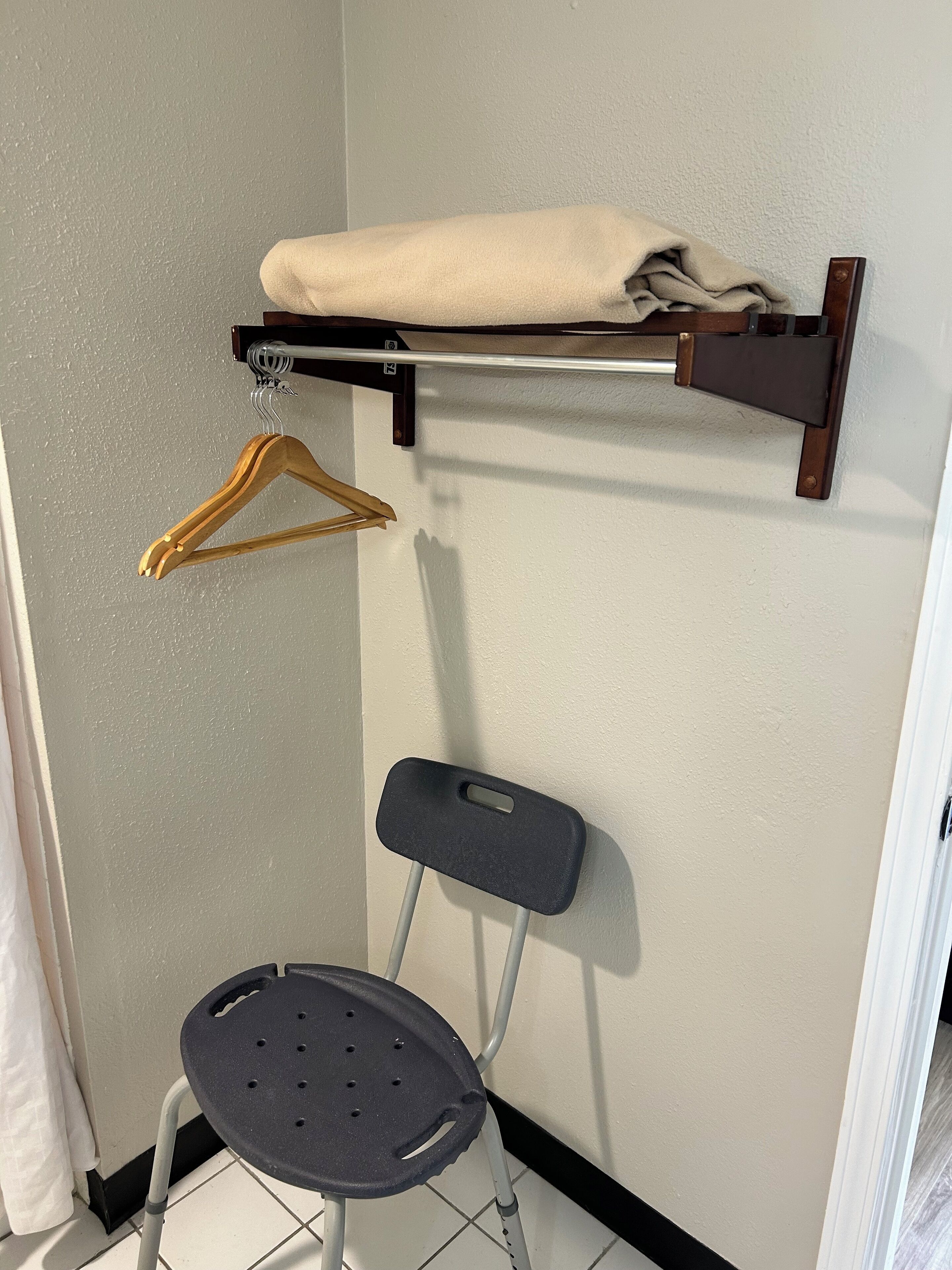 A corner of a room with a wall-mounted shelf holding a folded towel, a wooden hanger, and a black chair with a tray.