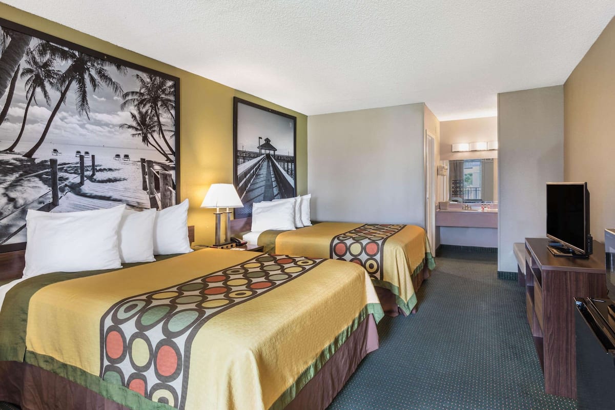 A hotel room with two beds, a desk with a TV, a bathroom with a sink and mirror, and a large beach mural on the wall.