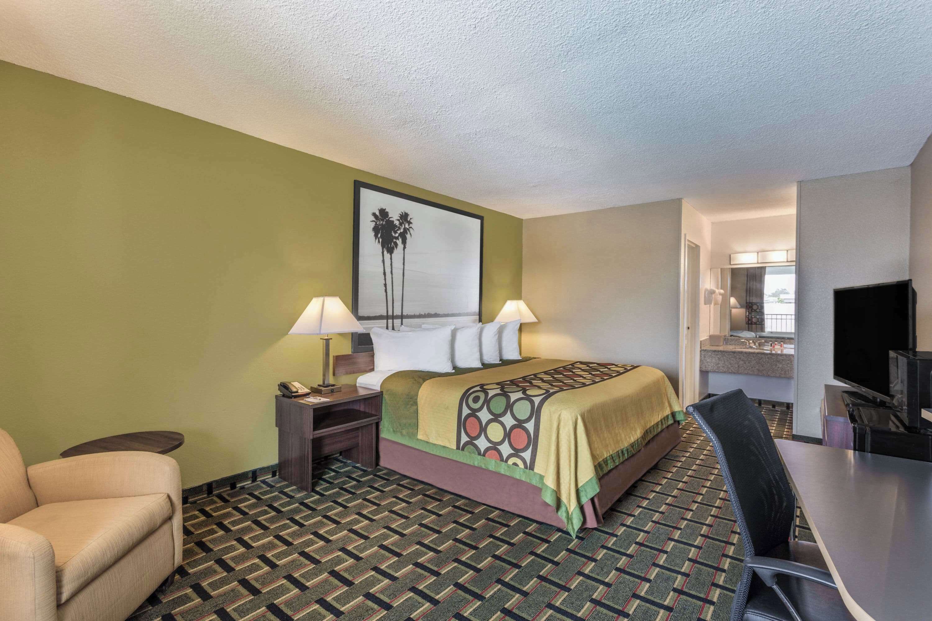 A hotel room with a bed, two bedside lamps, a desk with a chair, a television, and a bathroom visible through an open door.