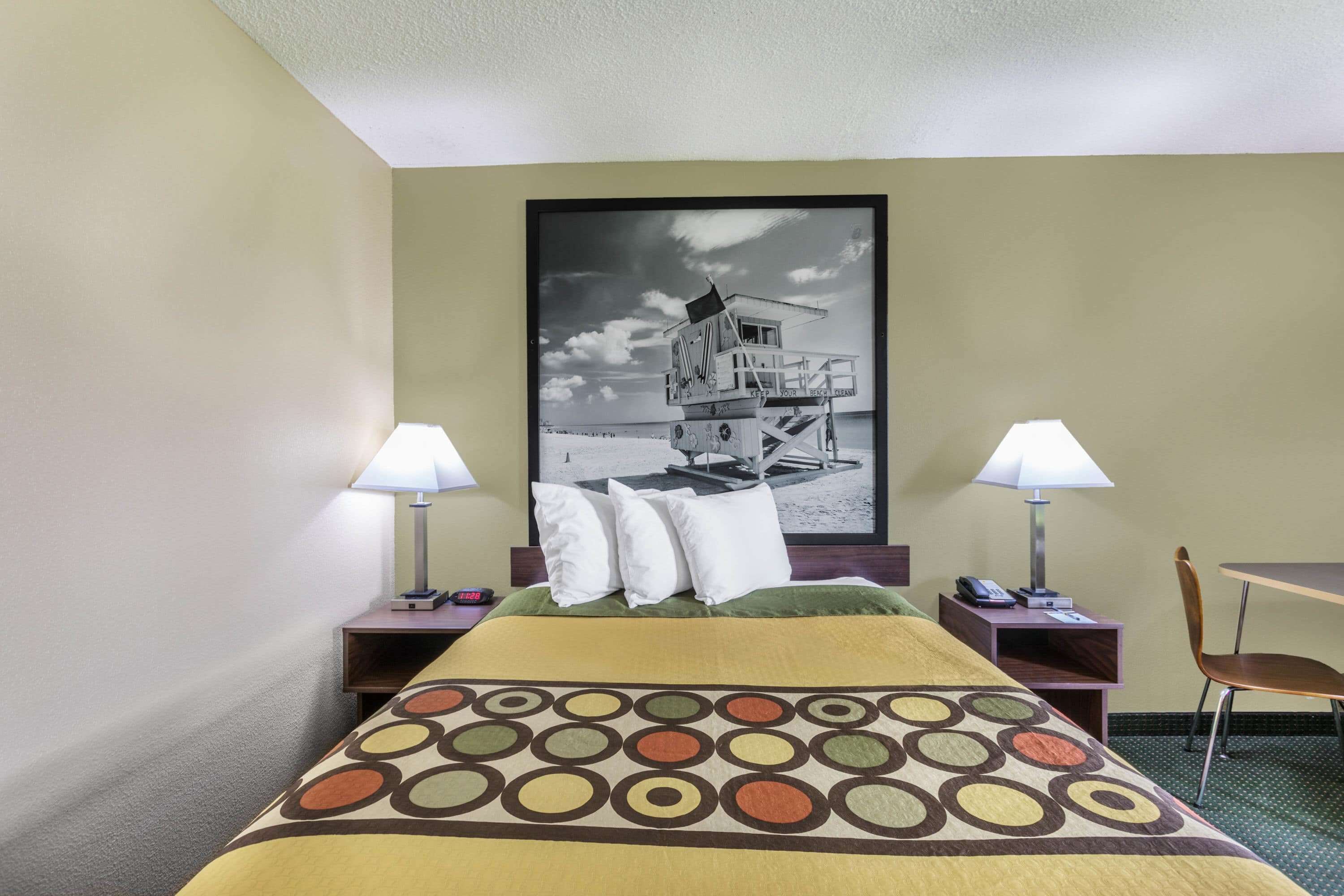 Une chambre d’hôtel comprenant un lit, deux tables de chevet, une chaise, un bureau et un grand tableau représentant un phare, accroché au mur.