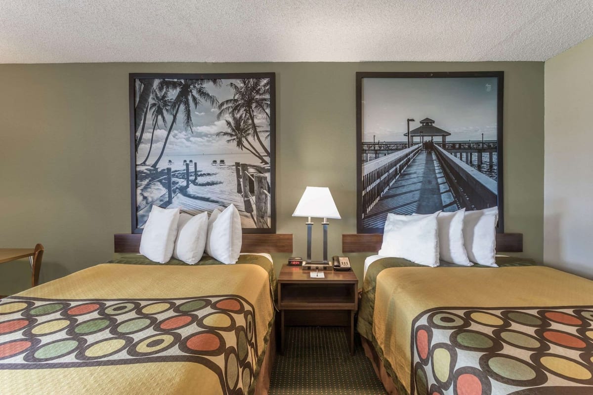 A hotel room with two beds, a nightstand, a lamp, and two large wall art pieces depicting coastal scenes.