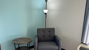 A corner room with a grey armchair, a small round table, a floor lamp, and a wall-mounted air conditioner.