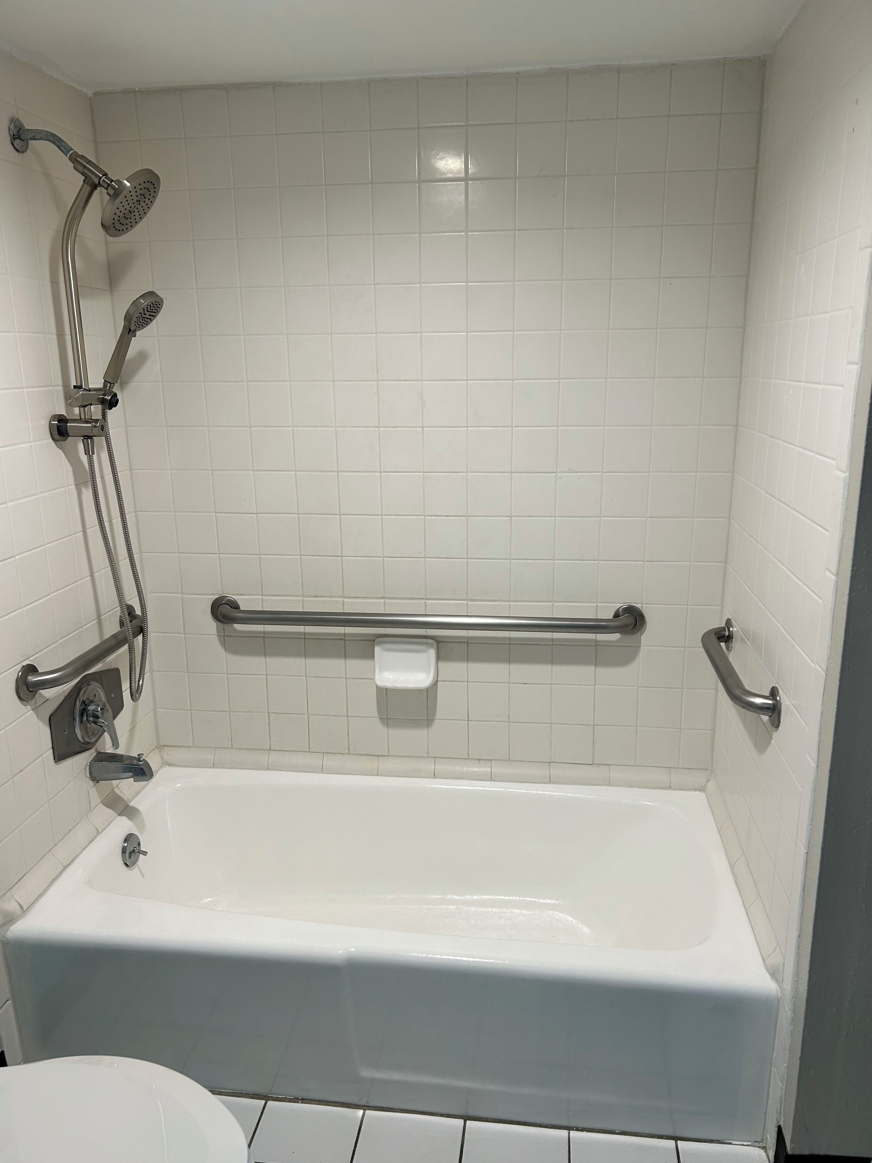 A white-tiled bathroom with a bathtub, a handheld showerhead, a handheld showerhead, a grab bar, and a soap dish.