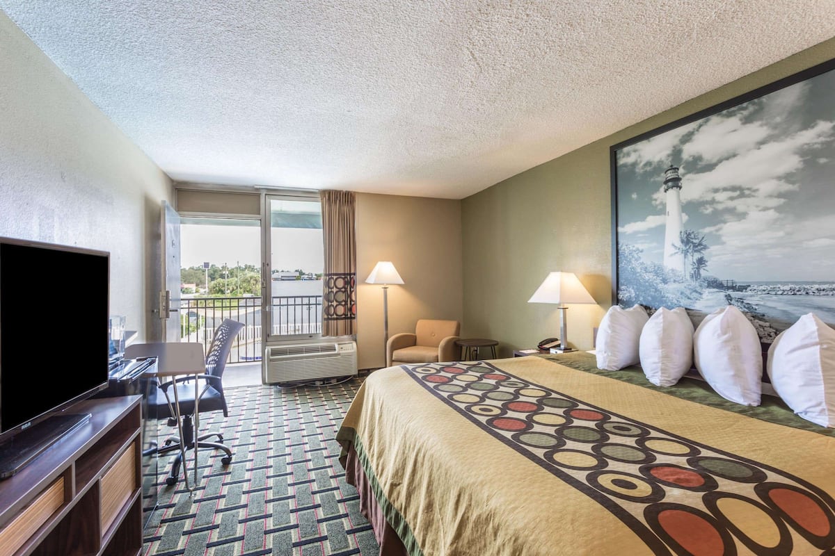 A hotel room with a large bed, a desk with a flat-screen TV, a chair, a balcony with a view, and a painting of a lighthouse.