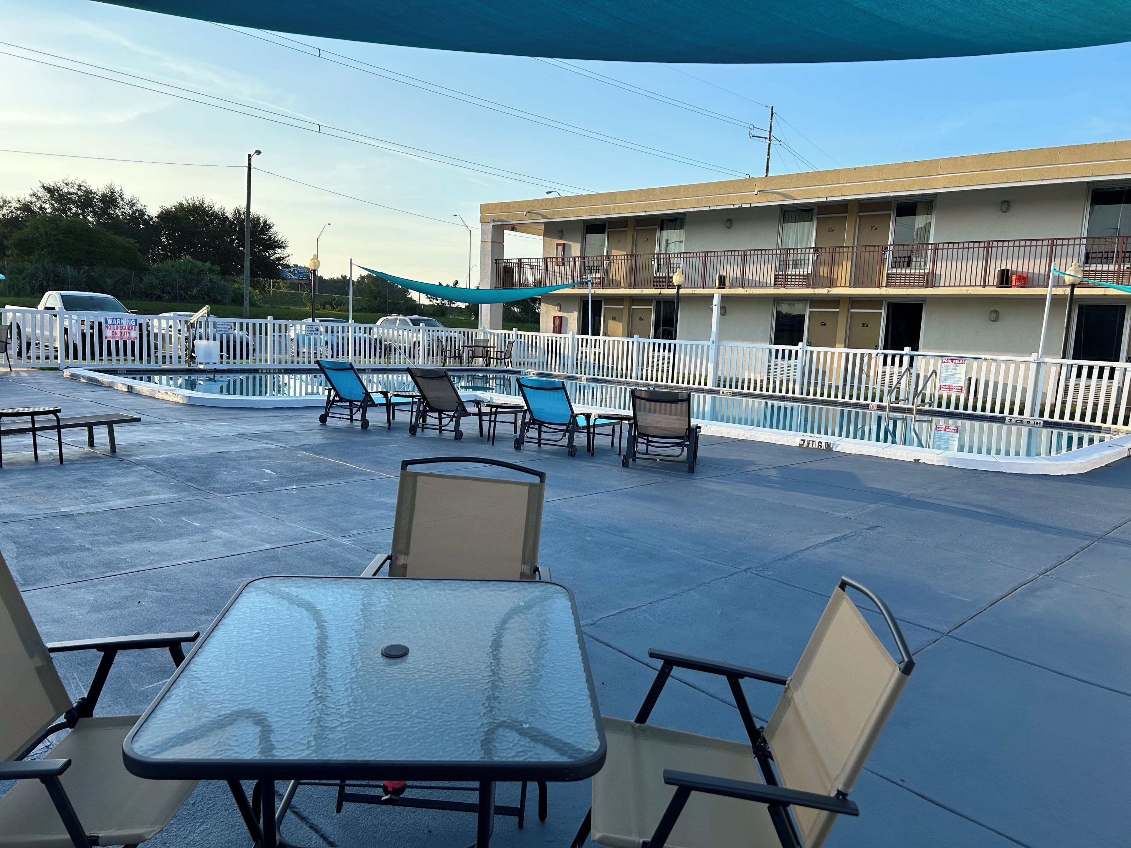Un área de piscina al aire libre con sillas de descanso y una mesa, rodeada por una valla blanca.