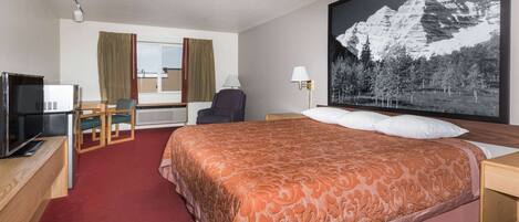 Habitación de hotel con cama, escritorio, silla, televisor y una fotografía en grande de montañas.