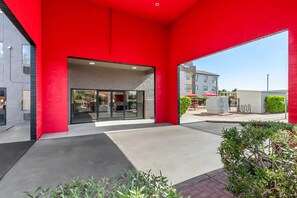 Een modern gebouw met een rode gevel, een glazen entree en een bestraat buitengebied met beplanting.