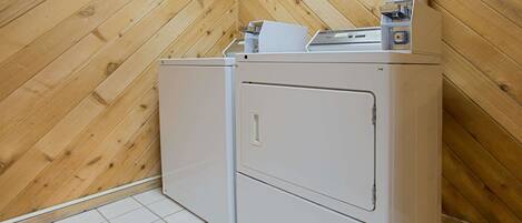 A laundry room with two white appliances, a wooden sloped ceiling, and white tiled flooring.