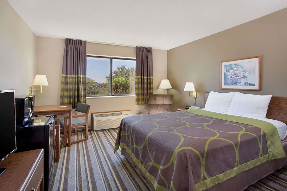 A hotel room with a bed, desk, chair, TV, lamps, and a painting on the wall.
