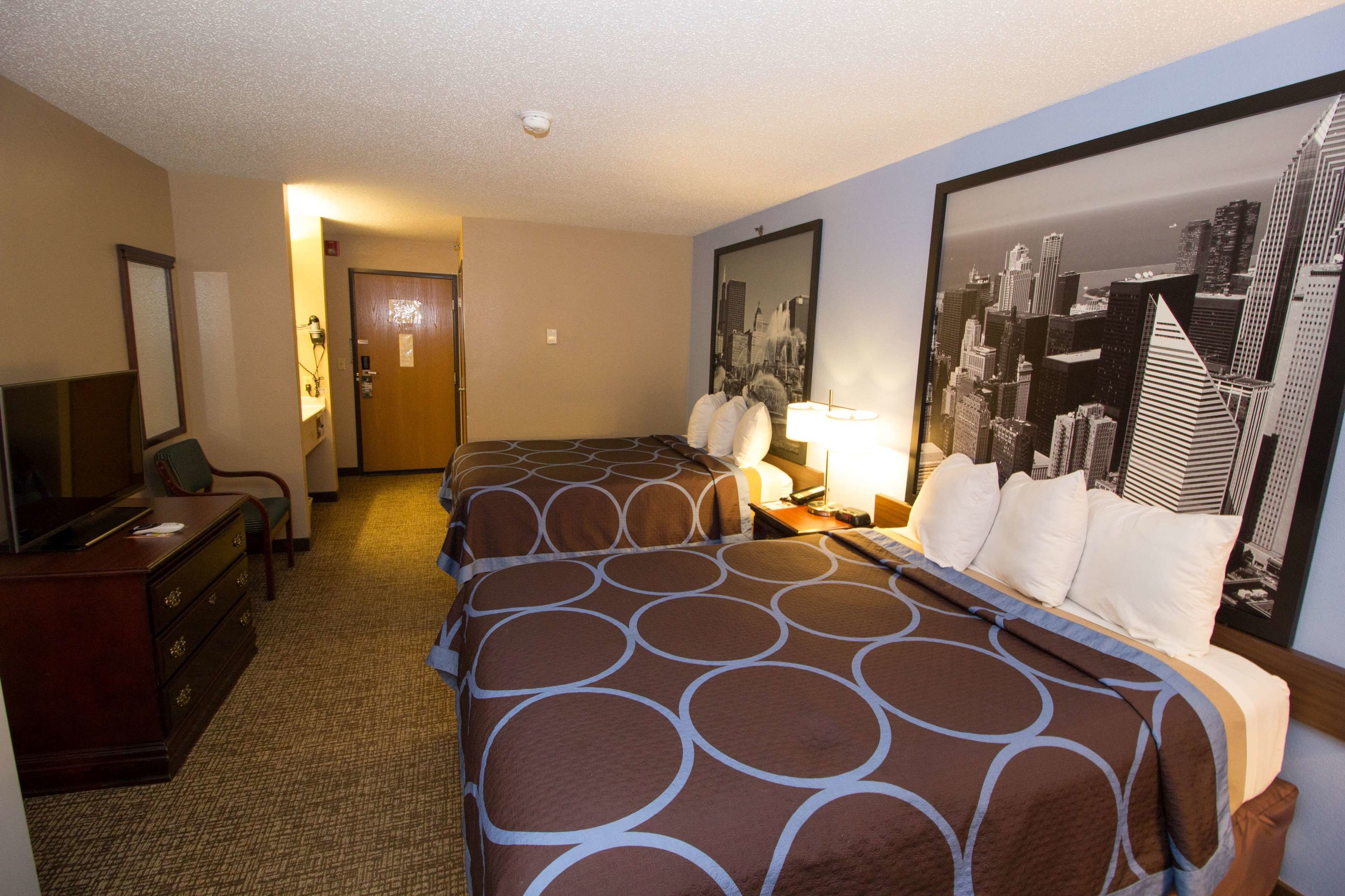 A hotel room with two beds, a desk with a computer, a chair, a dresser, and a cityscape painting on the wall.