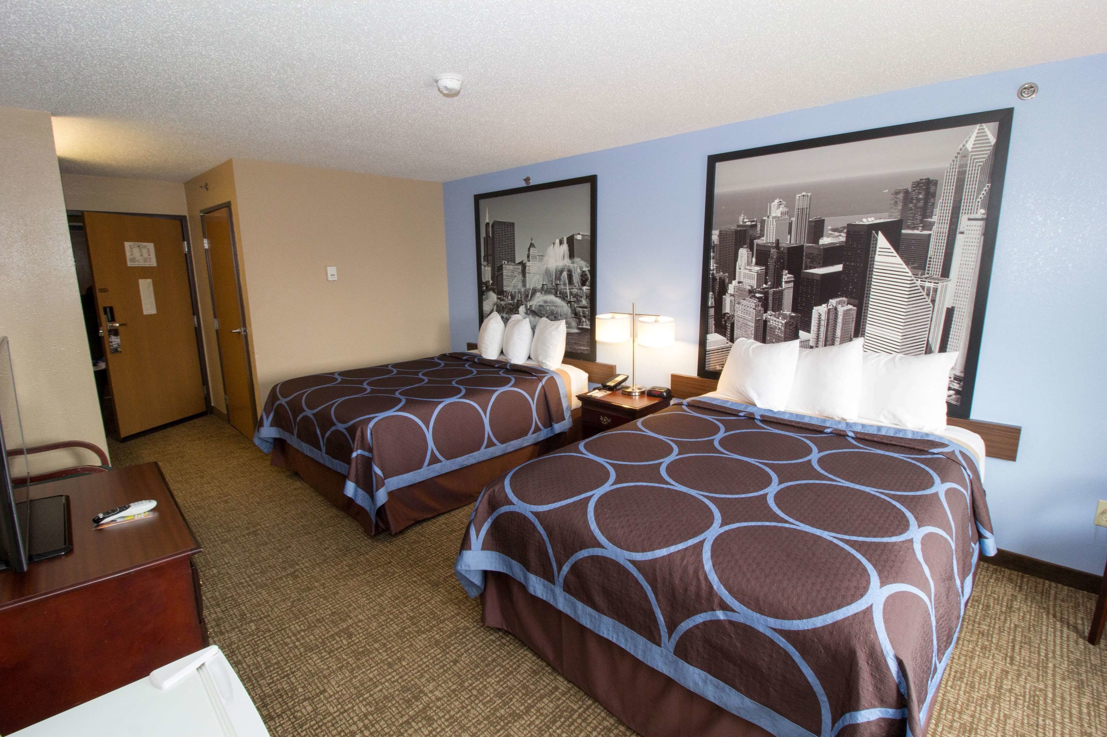 A hotel room with two beds, a desk, and a chair. There is a cityscape painting on the wall.