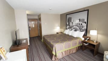Habitación de hotel con una cama grande, un escritorio con computadora, una silla, una mesita de noche con lámpara y una fotografía enmarcada de un paisaje urbano en la pared.