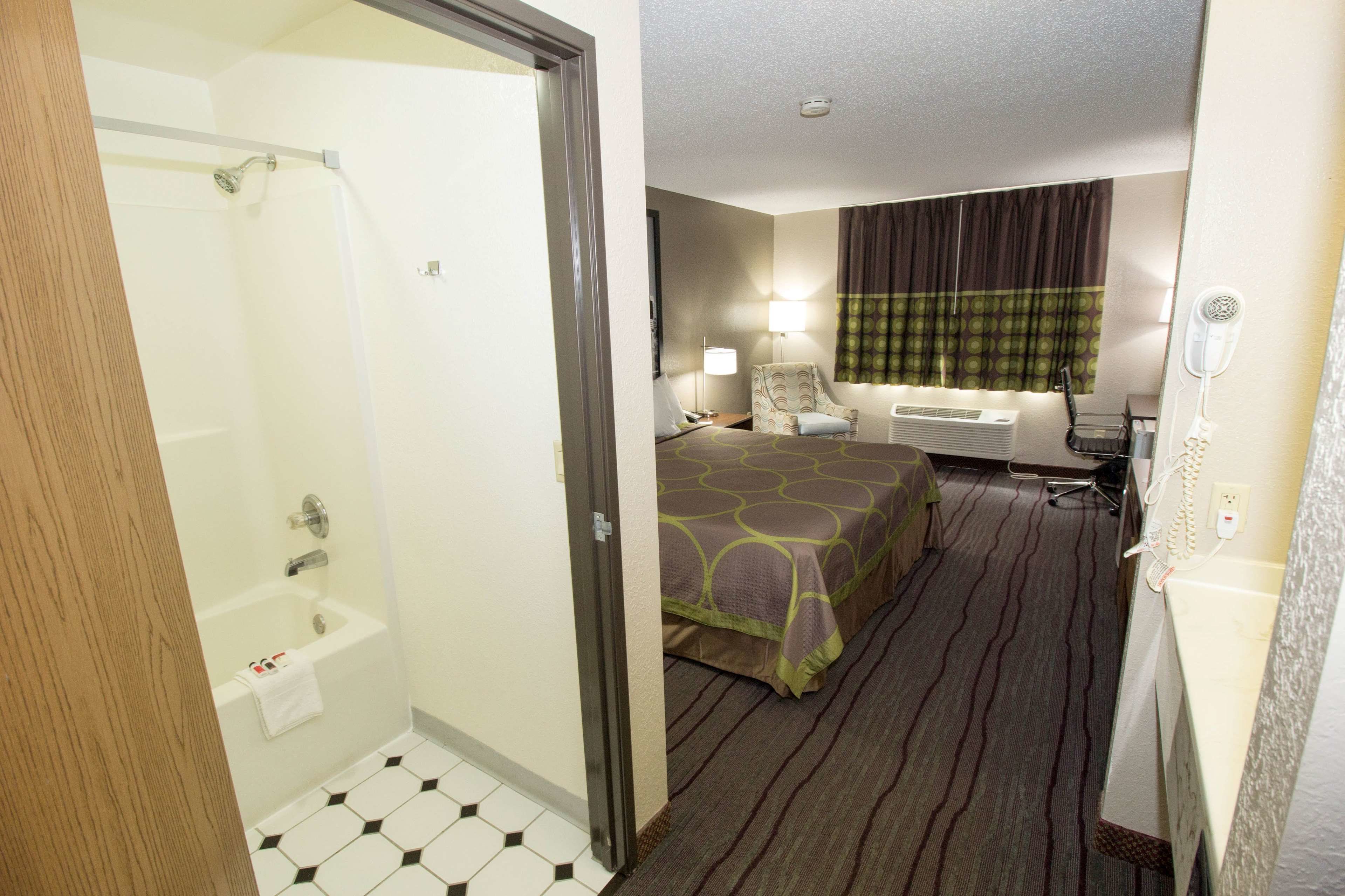 A hotel room with a shower, a bed with a patterned bedspread, a chair, a desk with a lamp, and a hairdryer.