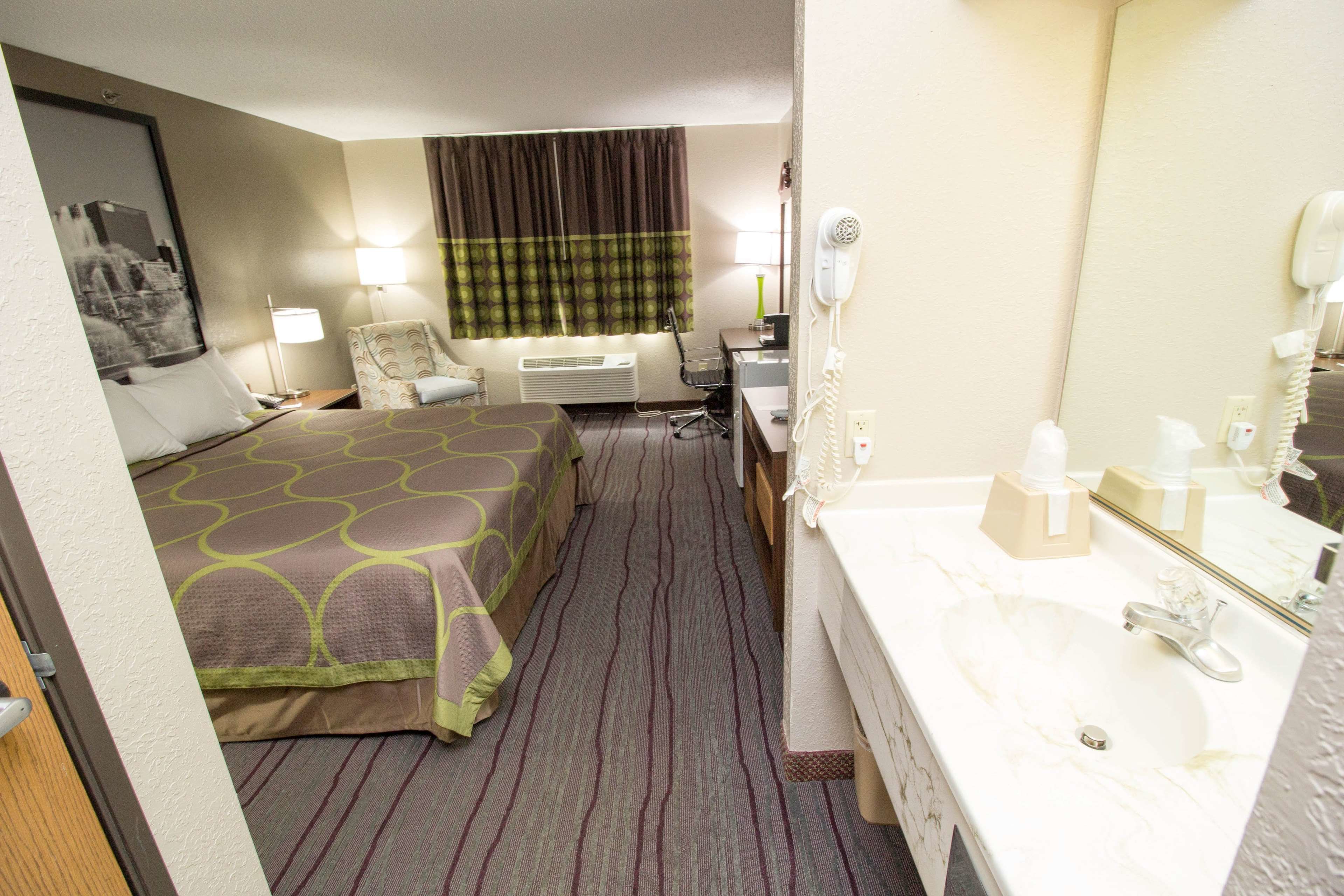A hotel room with a bed, a desk with a chair, a desk lamp, a hairdryer, a telephone, and a bathroom with a sink and mirror.