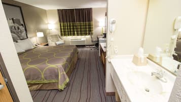 A hotel room with a bed, a desk with a chair, a desk lamp, a hairdryer, a telephone, and a bathroom with a sink and mirror.