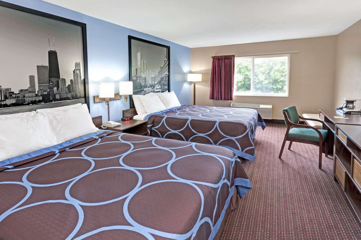 Ein Hotelzimmer mit zwei Betten, einem Schreibtisch, einem Stuhl, einem Fenster mit Vorhängen und einem Stadtbild an der Wand.