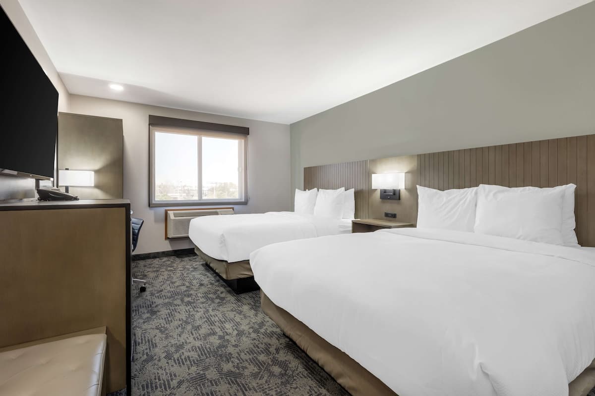 Ein Hotelzimmer mit zwei Betten, einem Schreibtisch, einem Fernseher und einem Fenster mit Blick auf die Stadt.