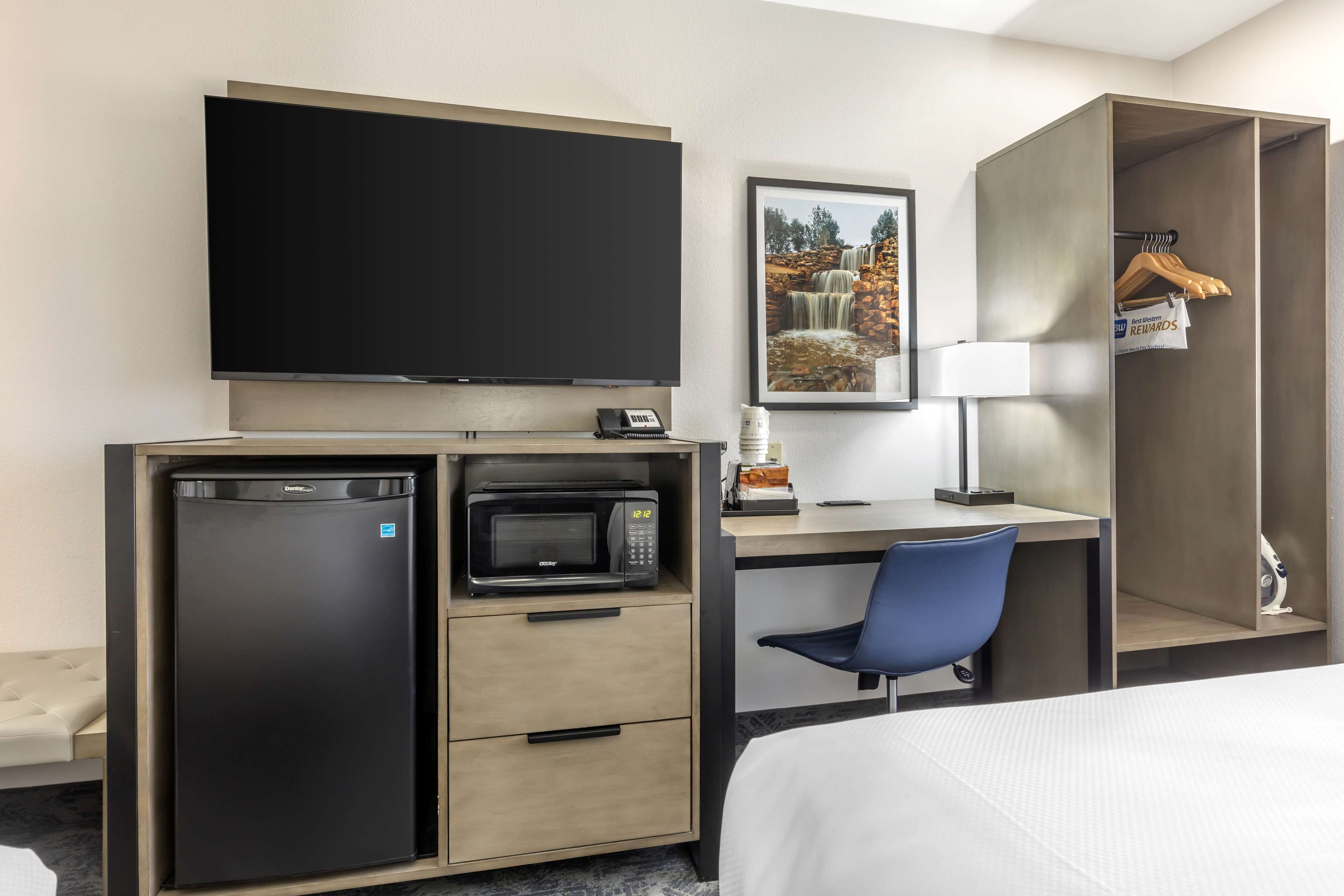 A hotel room with a bed, a desk with a chair, a television, a microwave, a refrigerator, a wardrobe, and a framed picture on the wall.