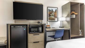 Une chambre d’hôtel comprenant un lit, un bureau avec une chaise, une télévision, un four à micro-ondes, un réfrigérateur, une armoire et un tableau encadré au mur.